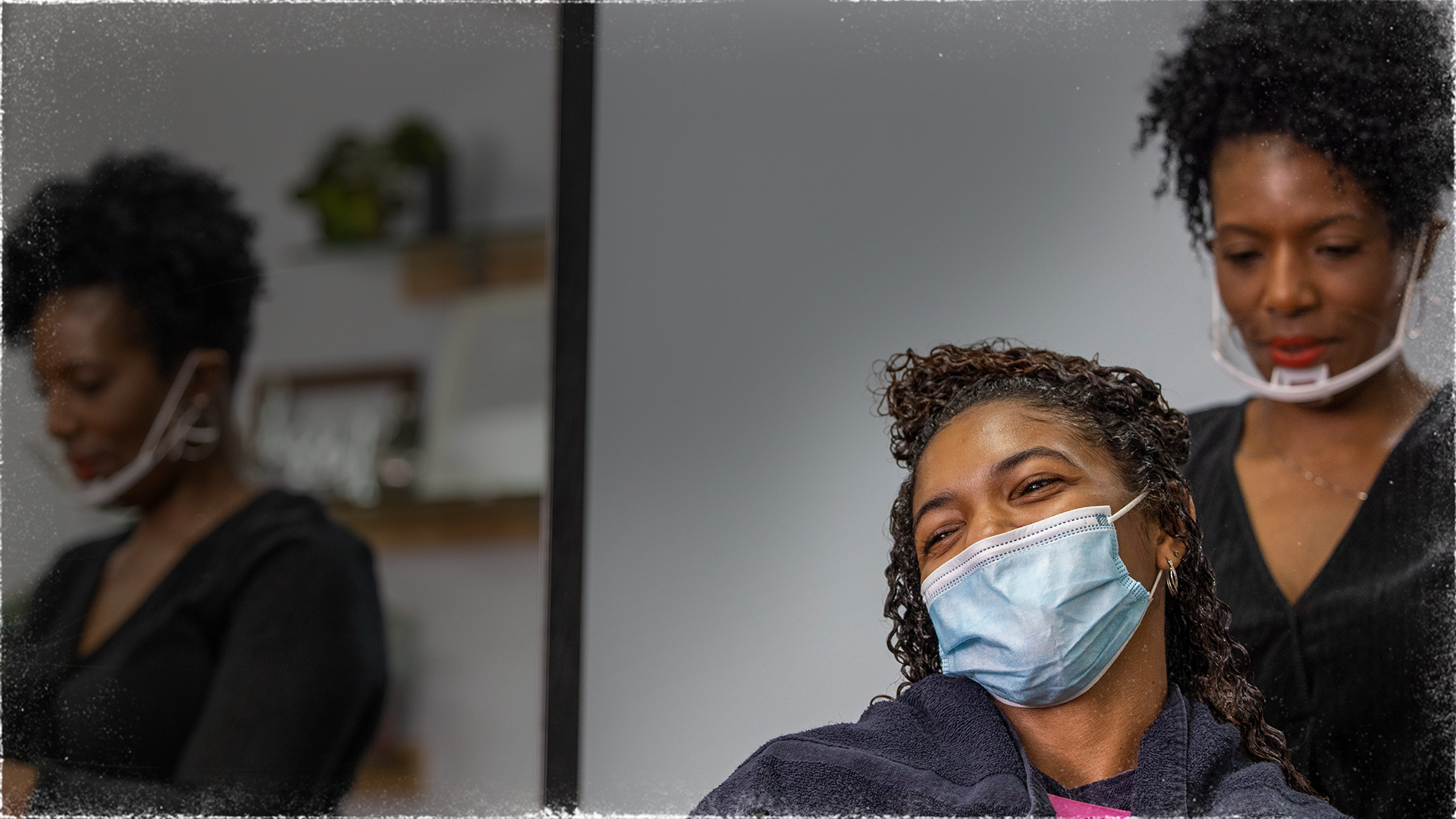 A woman wears a face mask in a stylist chair in a salon. Behind her, another woman wearing a mask is styling the first woman's hair. 