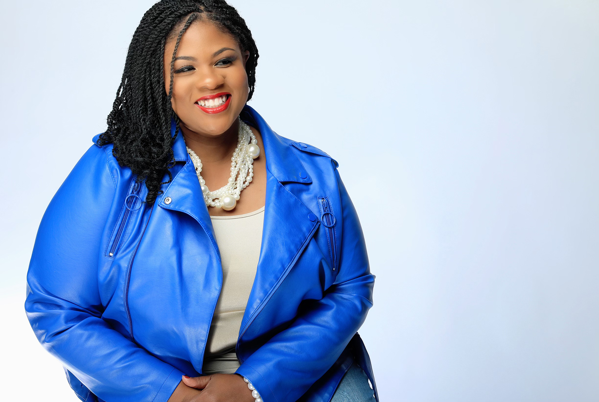 An image of a woman sitting on a chair, smiling for a photo against a white background while wearing a bright blue blazer.