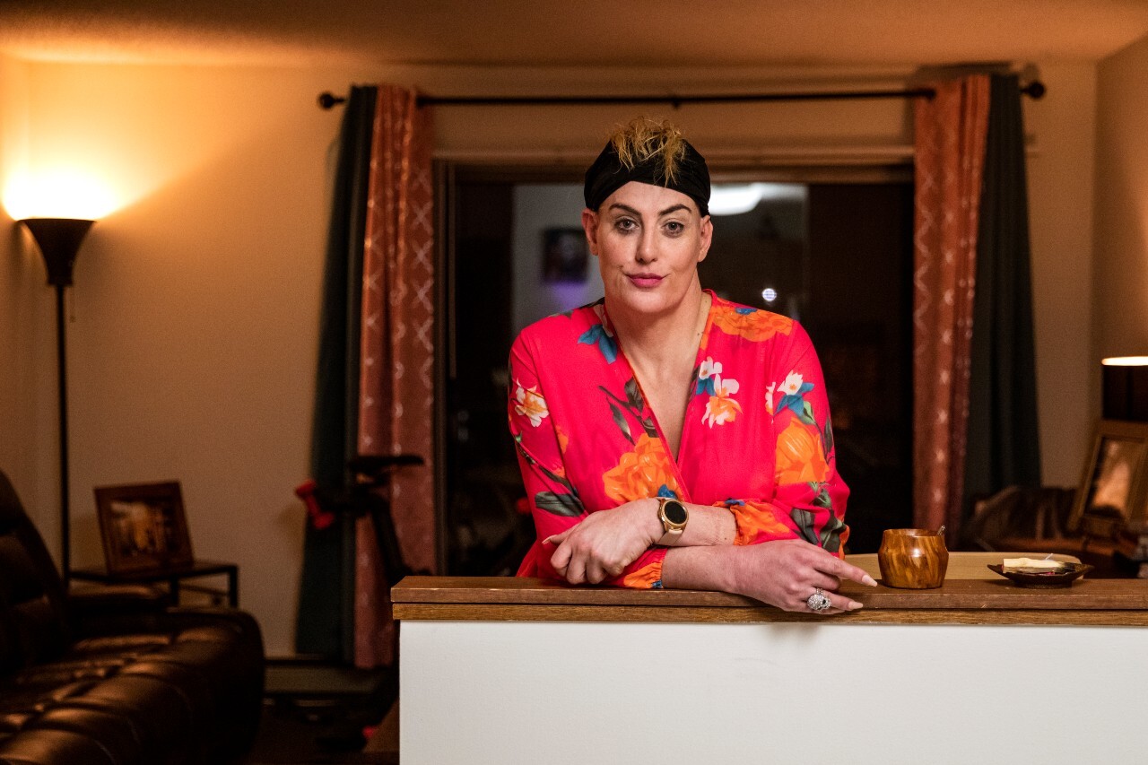 An image of a woman softly smiling for a photo while standing at the kitchen counter in her apartment. She is wearing a black headband and a pink floral robe.