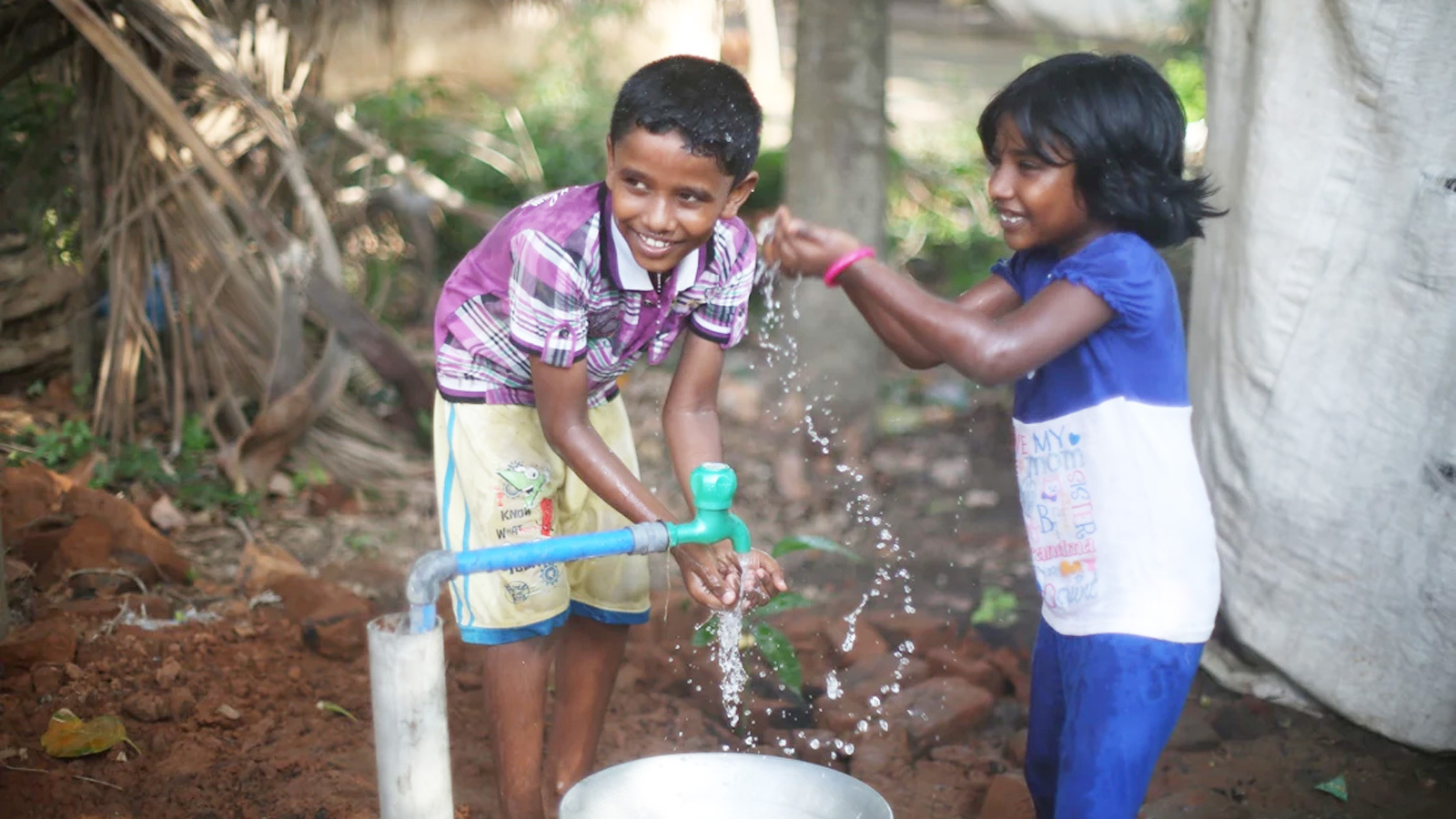 Two children smiling while pumping water from a well in a rural village