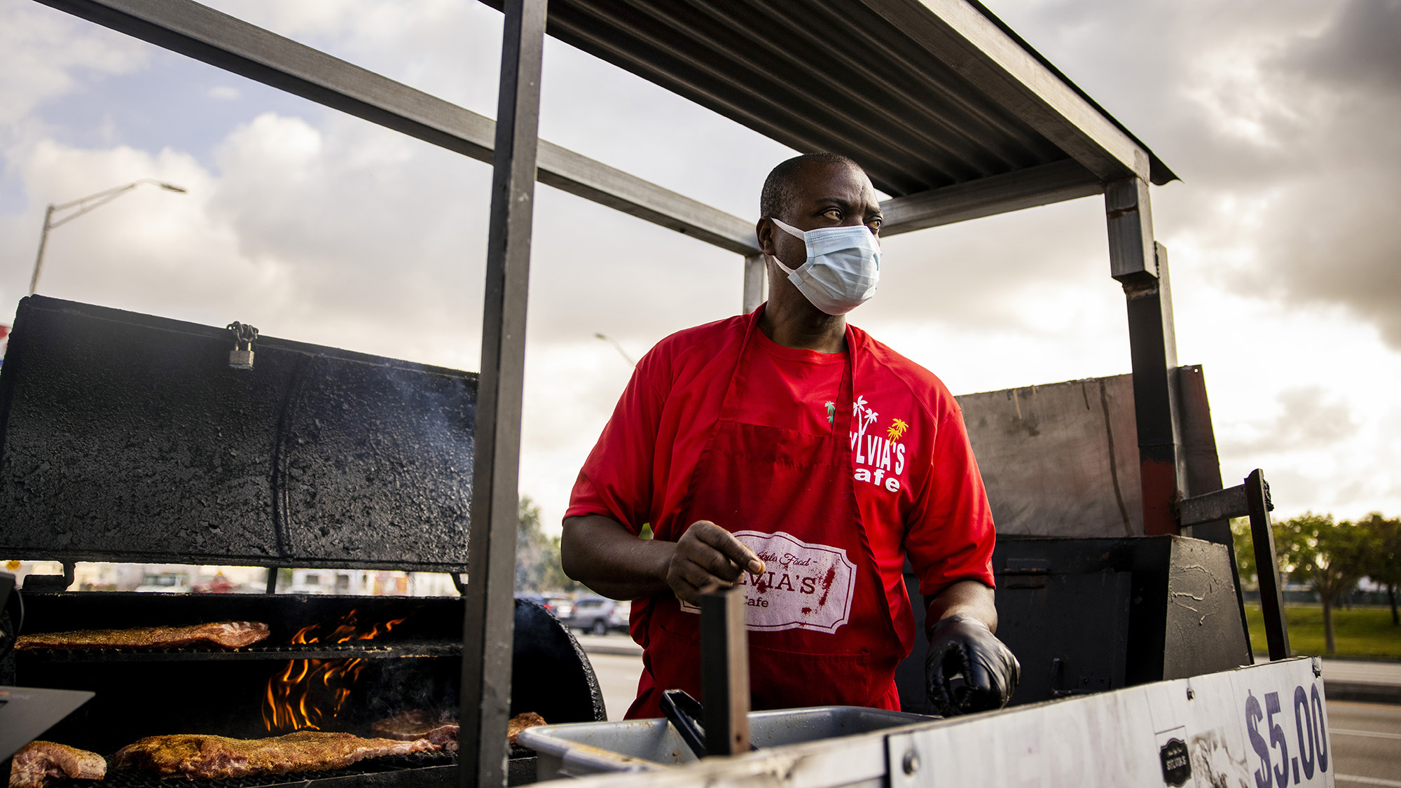 A man stands in front of an outdoor smoker. He wears a mask and holds products to prepare meals for customers. 