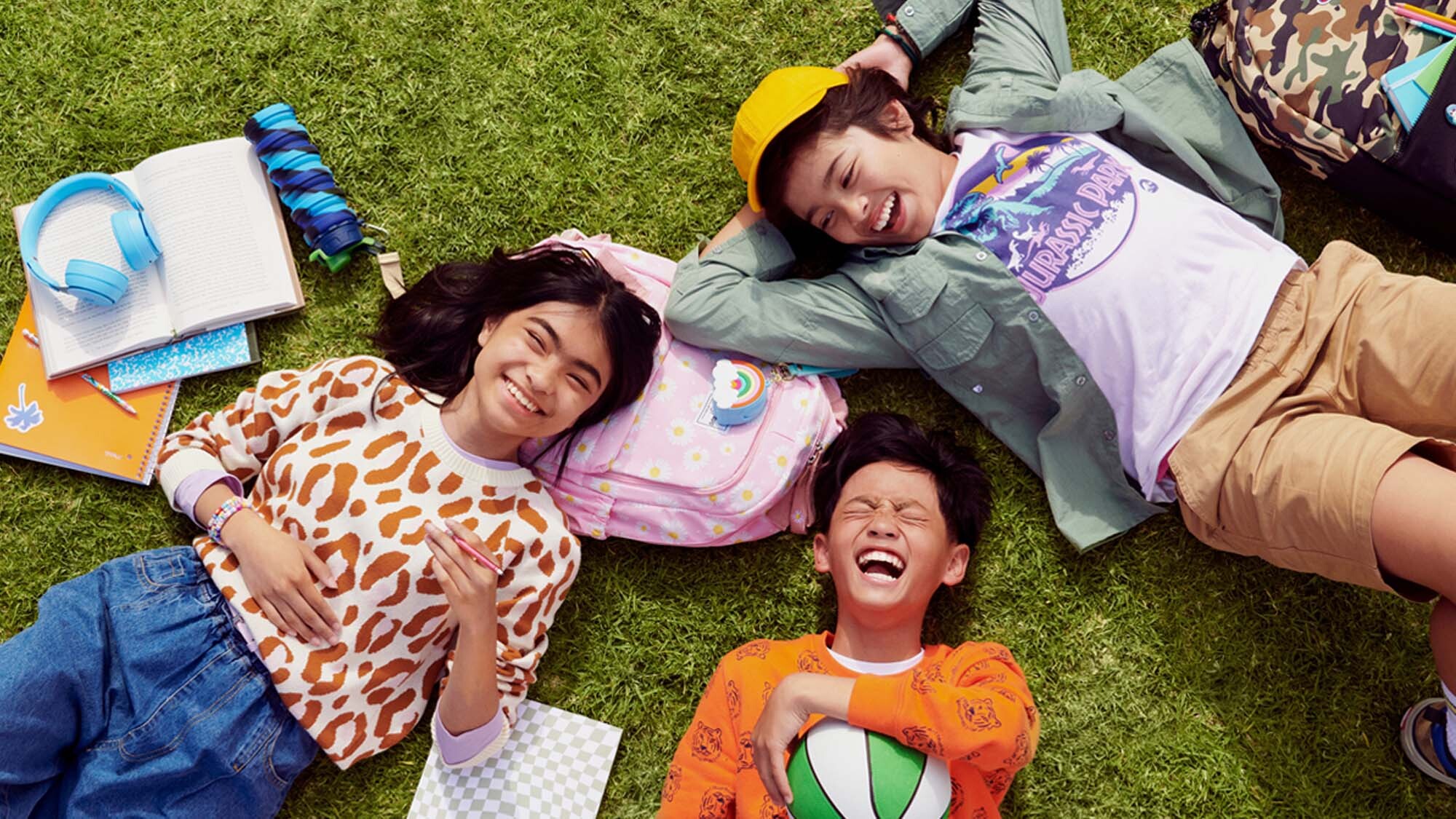 Image of three kids lying on grass with school supplies scattered around them.