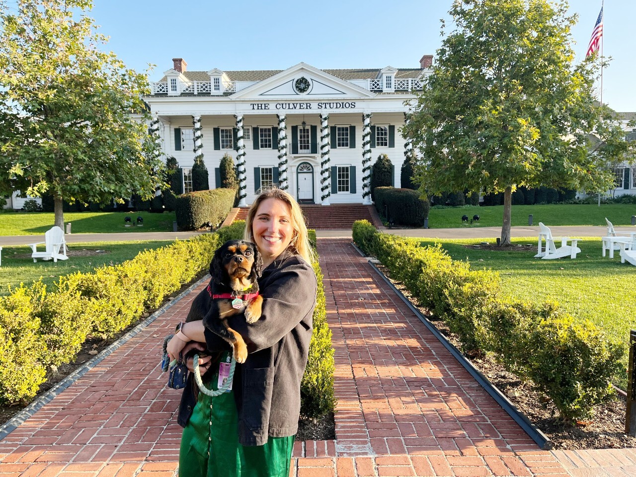 An image of a woman holding a dog and smiling while standing in front of a large white colonial style building that says "The Culver Studios" on it.