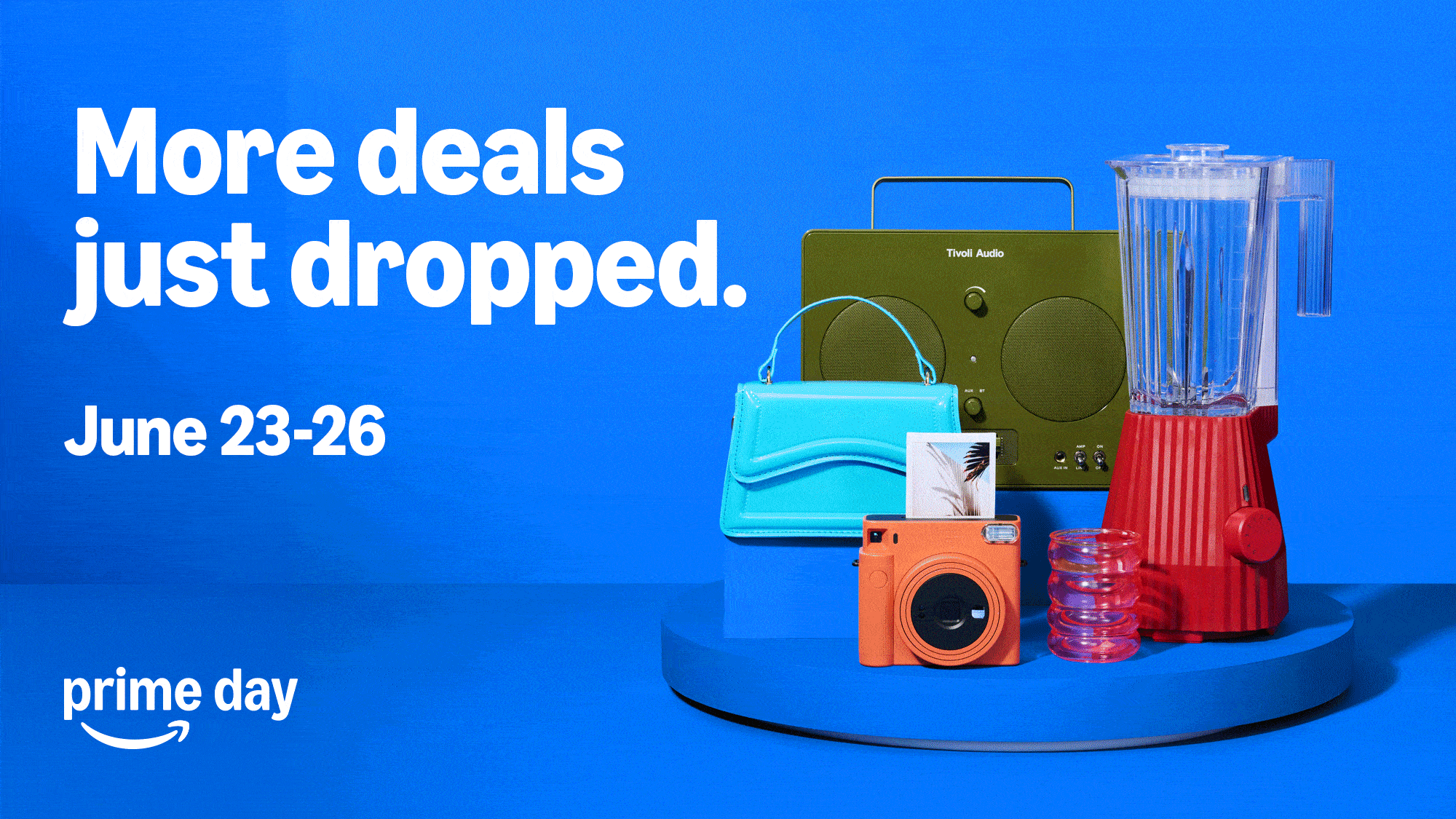 White text over a blue background reads: "More deals just dropped. Amazon Prime Day 2026 is June 23-26." A green speaker, blue purse, orange camera, red blender, and a spiraled red glass are staged on a shelf.