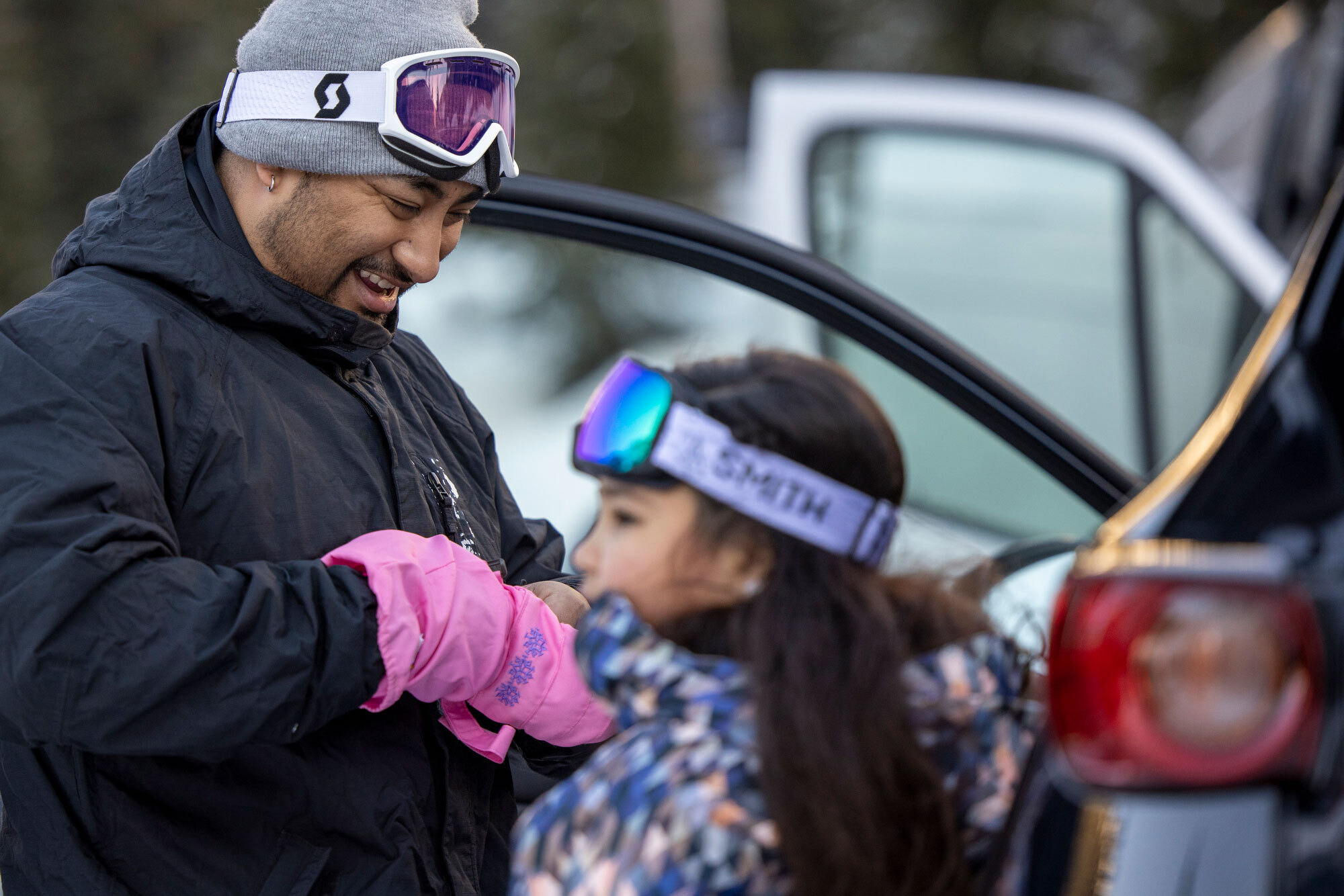 A father smiles as he looks down and helps his young daughter get her winter glove on.