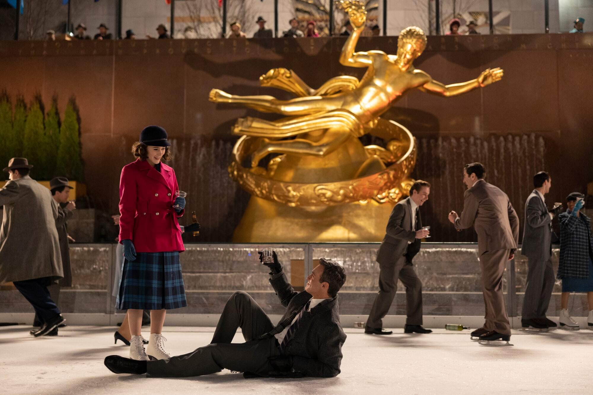 An image of Midge Maisel standing on an ice rink with a man on the ground below her reaching for her hand.