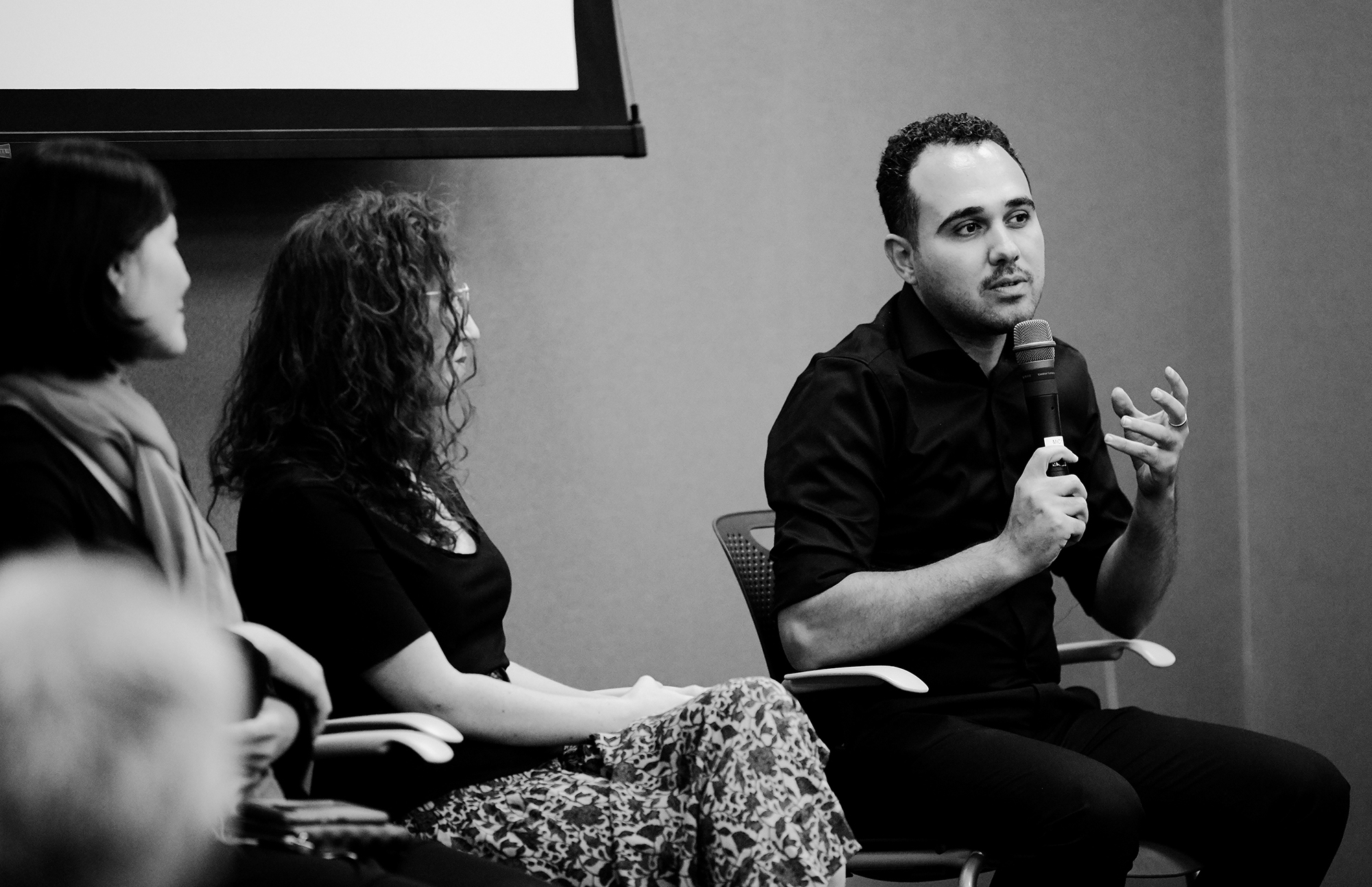 A man sits in a chair and talks into a microphone as part of a speaking panel with two females to the left.