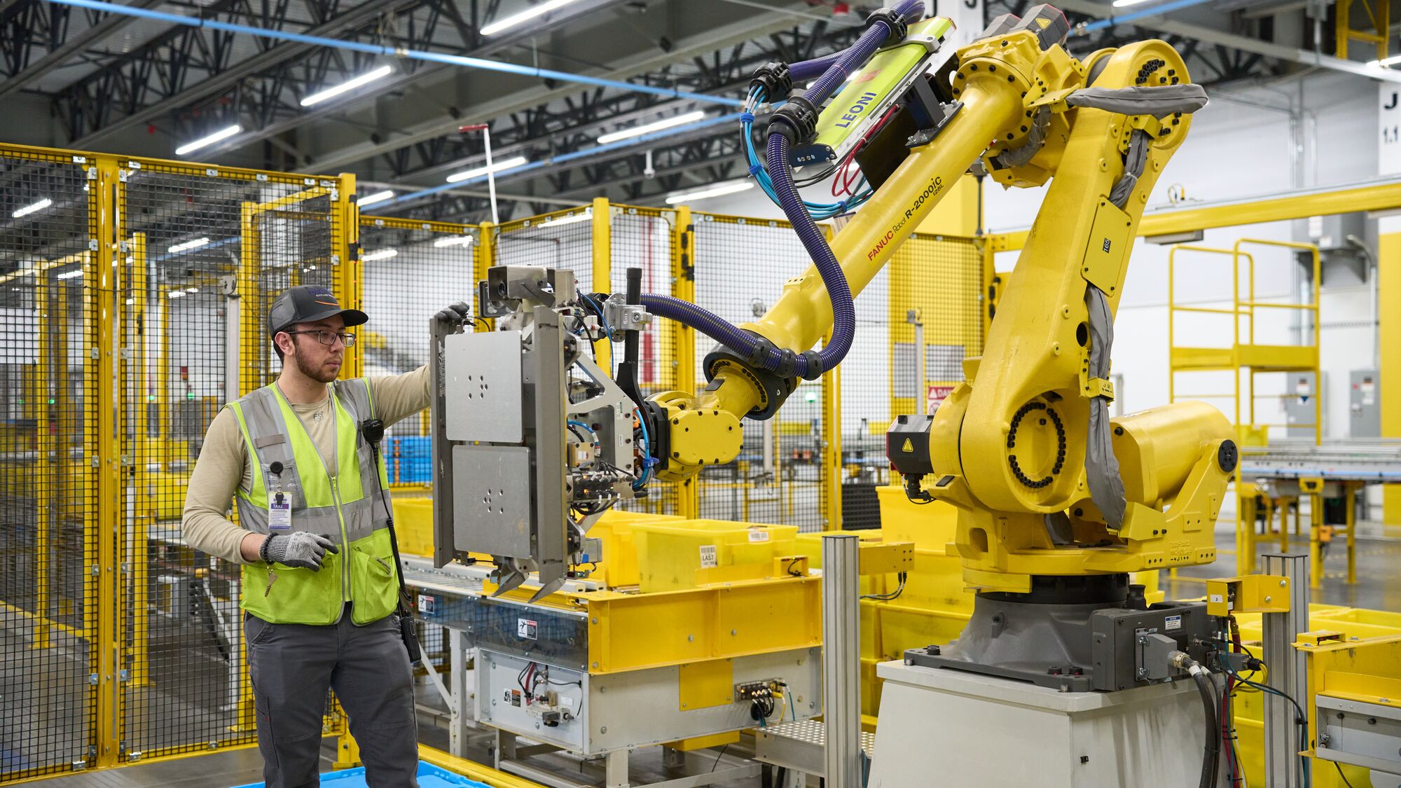 An Amazon employee in safety vest interacting with robotic equipment at a robotics facility